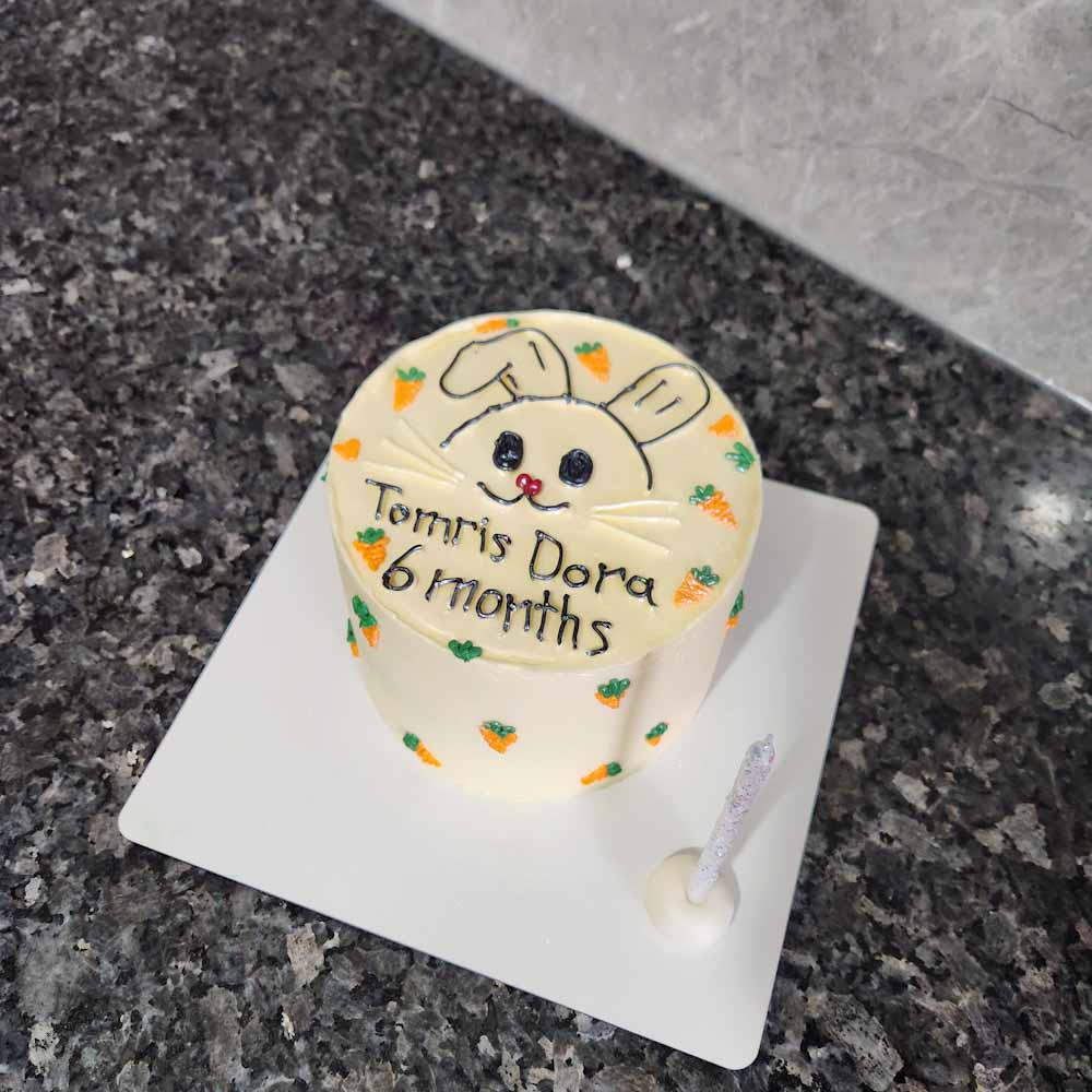 6 aylık bebek için hazırlanan butik pasta; üstünde sevimli tavşan çizimi ve etrafında minik havuç süslemeleriyle tasarlanmış. Pastel tonlarda, zarif ve eğlenceli bir görünüm sunuyor.