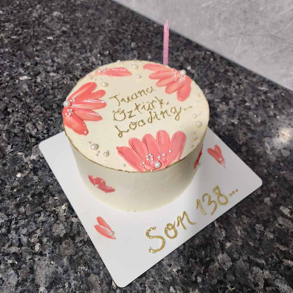 Yuvarlak butik pasta; pembe çiçek desenleri ve inci süslemeleriyle zarif bir şekilde hazırlanmış. Üzerinde altın renkle yazılmış ‘Tuana Öztürk Loading…’ ifadesi ve tek pembe mum yer alıyor. Pasta tablasında ise ‘Son 138…’ yazısı bulunuyor; kişiye özel, anlamlı bir kutlama tasarımı.