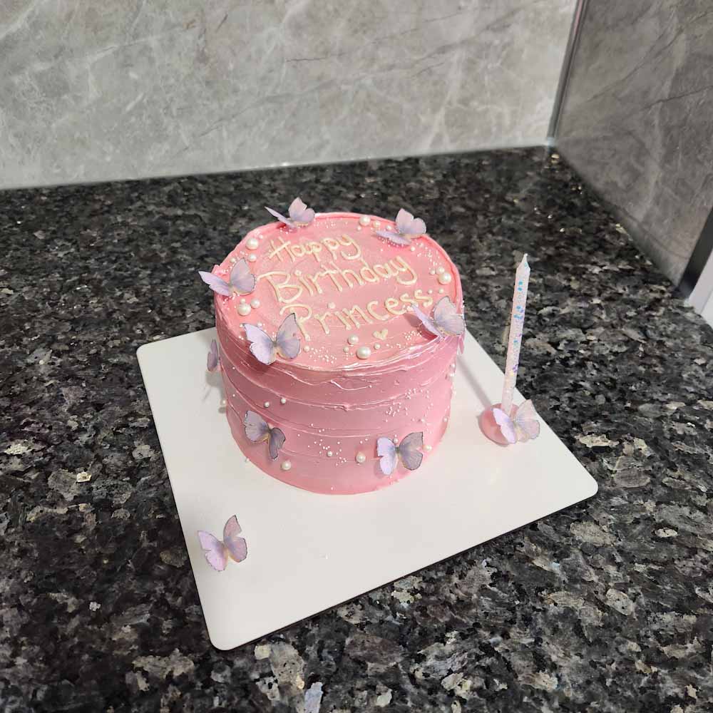 Zarif butik tasarım pasta; üstünde inci detaylarıyla süslenmiş beyaz zemin, kenarlarında pastel tonlarda kelebek figürleri. Üzerinde özel yazı ile kişiye özel hale getirilmiş şık doğum günü pastası.